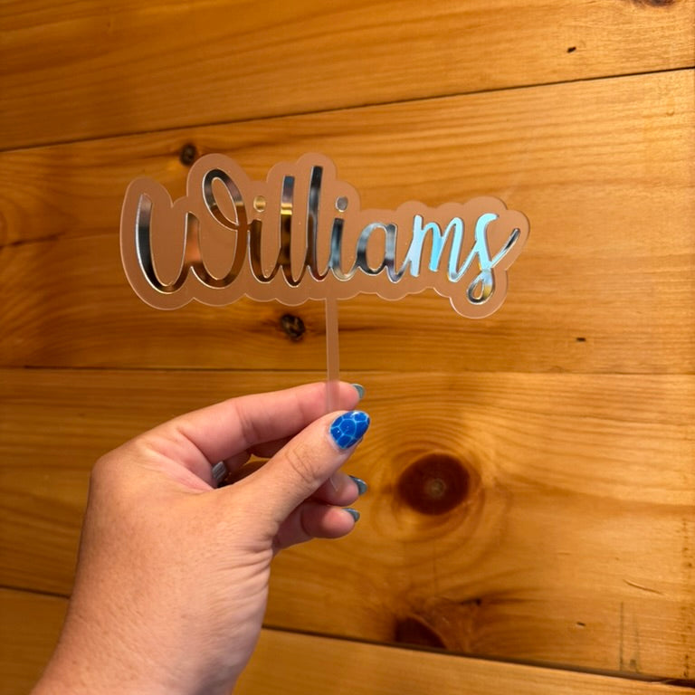 Hand holding a 'Williams' name sign against a wooden background